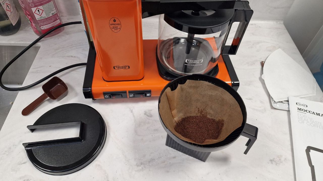 An orange moccamaster on a white countertop nest to a filter with used coffee grounds inside.