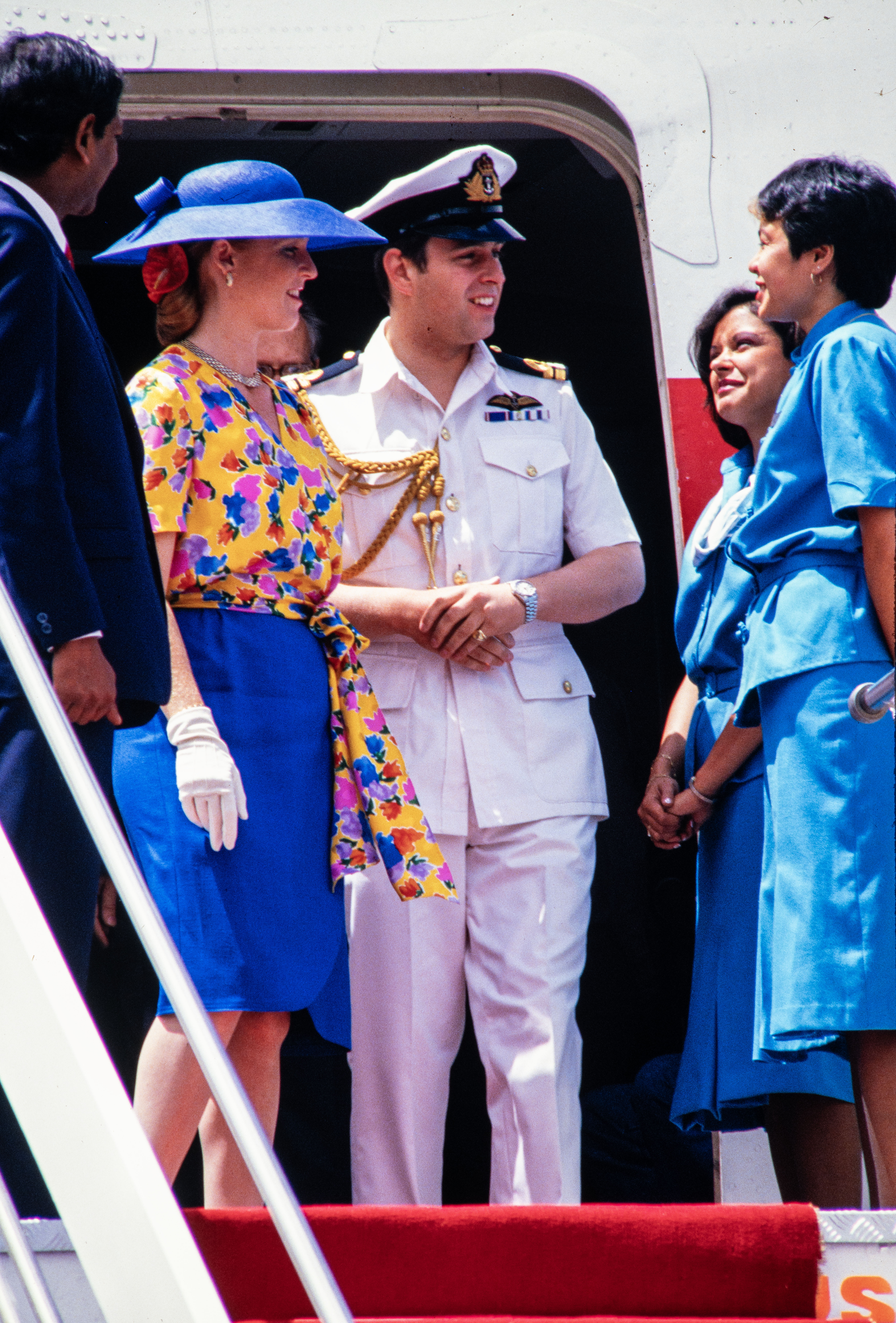 Sarah Ferguson wearing a blue skirt and hat and yellow floral top standing on the stairs of a plane next to Prince Andrew in a naval uniform