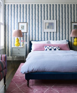 A pink and blue bedroom with white and blue striped wallpaper, pink rug and yellow lamps with pink fabric shades.