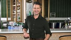Nate Berkus has shared many tips for personalizing your space. He is pictured wearing a button-down black shirt leaning on a bar with a white surface, with white and brushed gold shelves and green tiles in the background