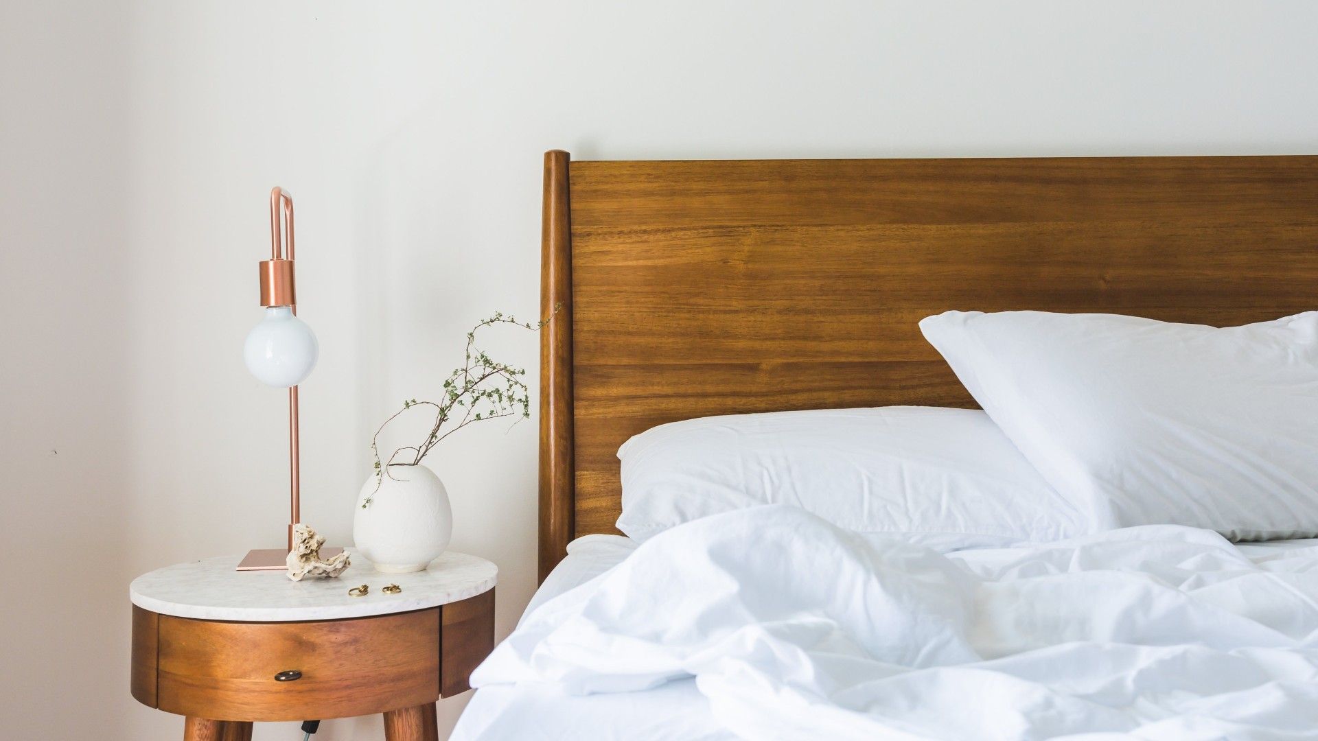 a clear marble-topped nightstand with flowers and lamp, next to a bed with a wooden headboard, and white painted walls