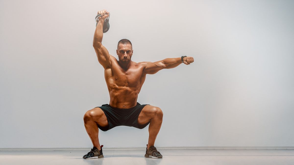 Man against grey backdrop performing a single arm overhead kettlebell squat with right arm raised