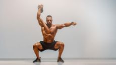 Man against grey backdrop performing a single arm overhead kettlebell squat with right arm raised