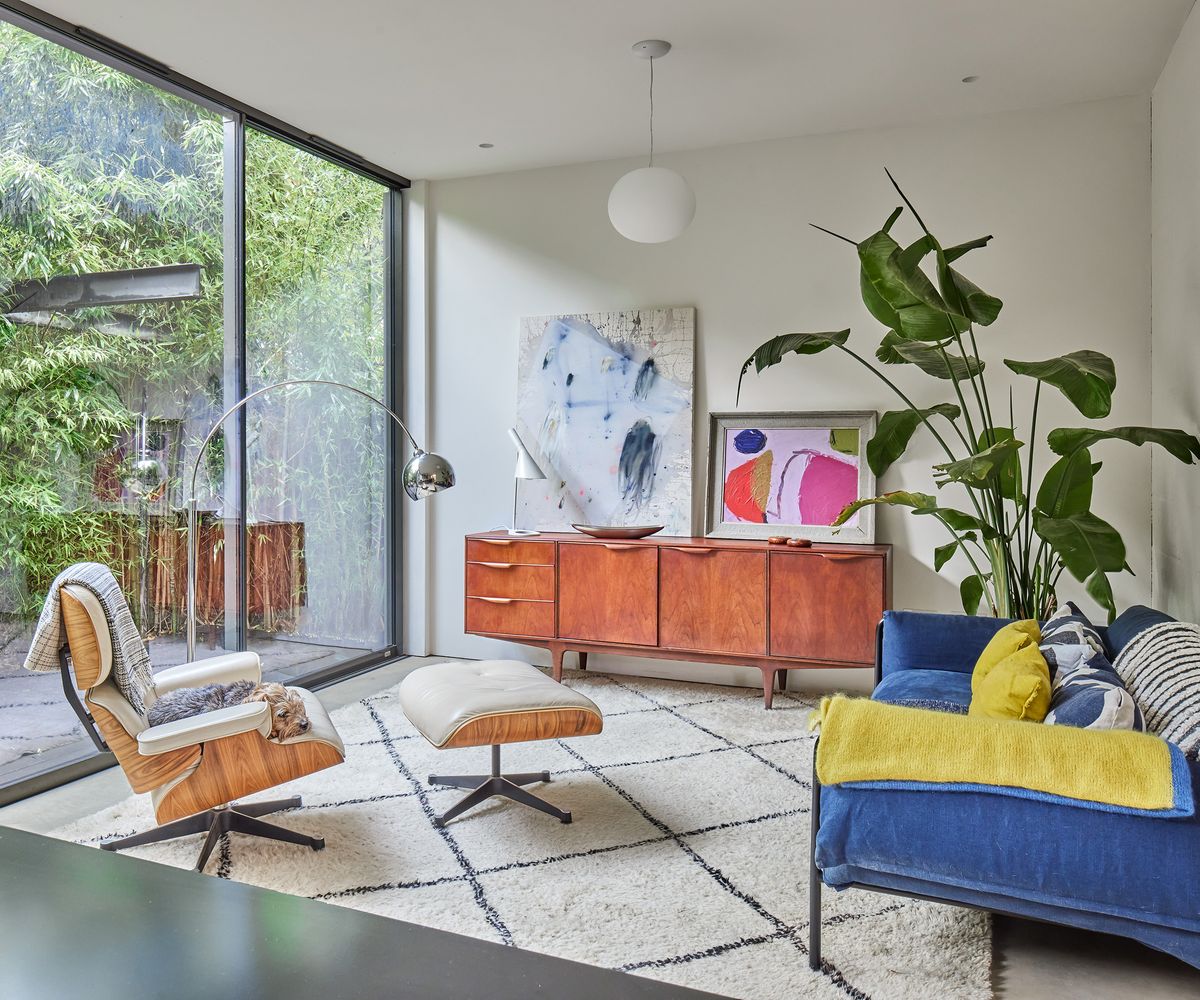 A modern living room space with a wall of glazing to the left looking out to the back garden. The furniture is mid-century modern style with a large statement potted houseplant and shaggy white rug with geometric pattern