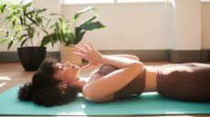 A woman lies on a yoga mat in her living room practicing yoga. She is on her back, with her eyes closed, and her hands clasped together in front of her chest in a prayer position. In the background are two leafy plants. 