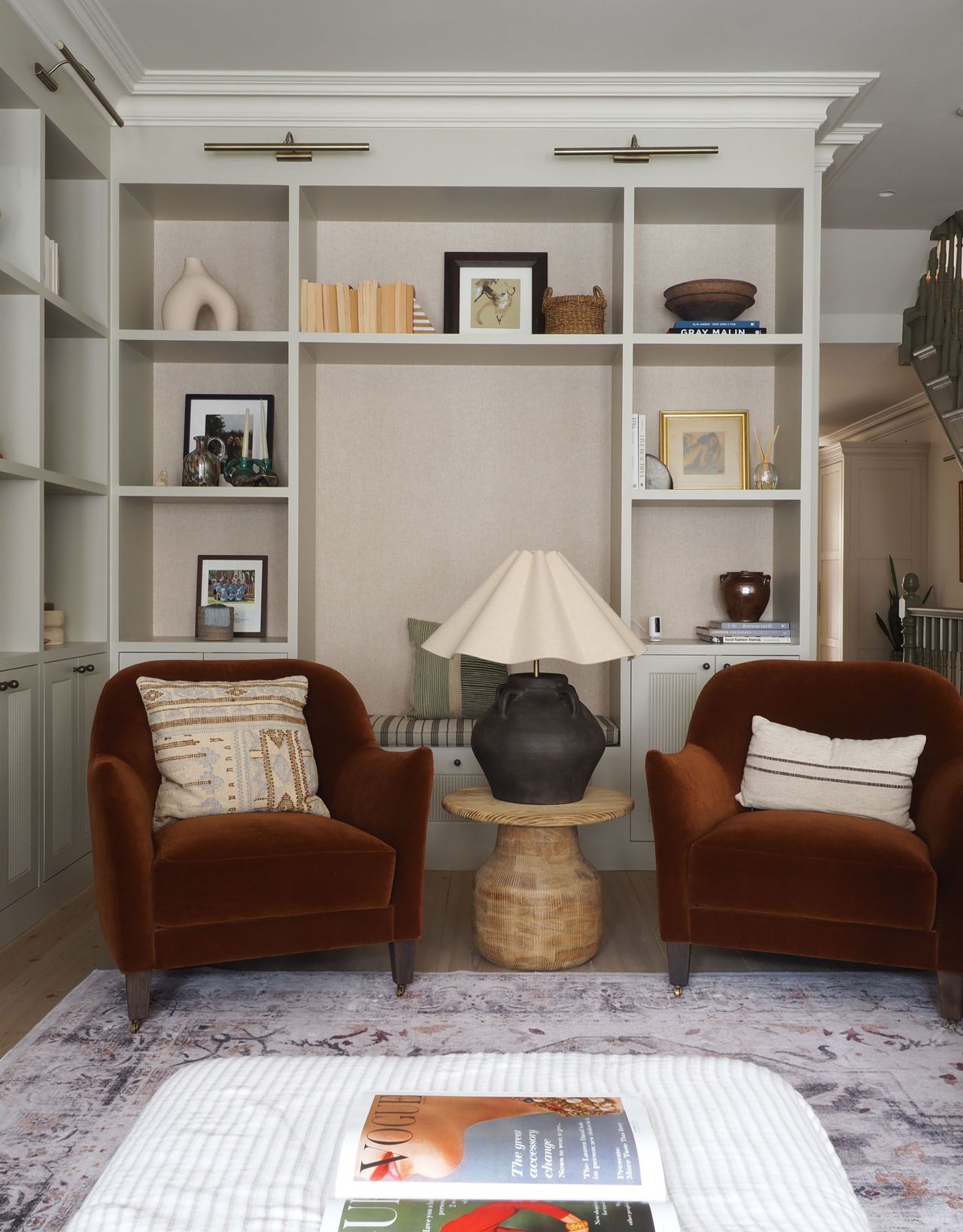A corner seating area with two rust velvet armchairs with beige cushions, a side table in the center with a large table lamp with a pleated shade in front of an alcove seat and decorative shelves