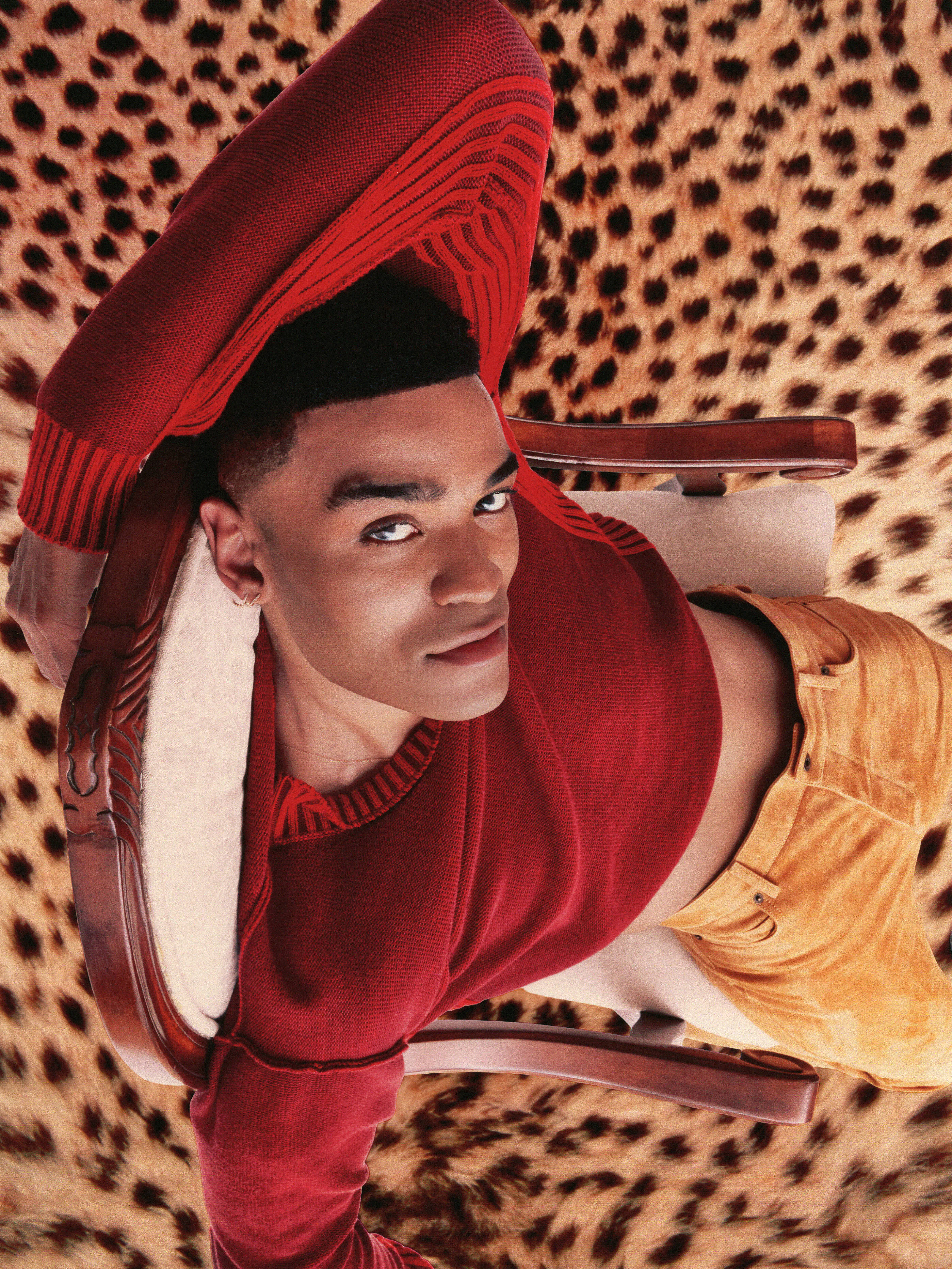 Owen Thiele poses in a chair over a leopard print rug wearing a red sweater and orange suede pants