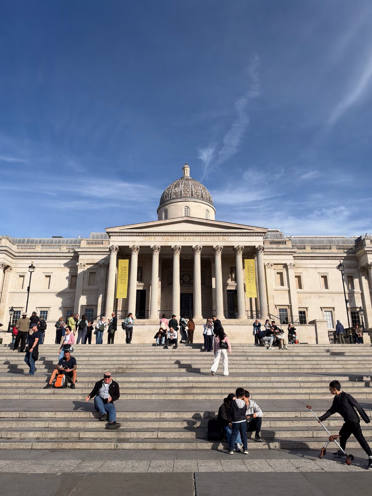 National Gallery in Trafalgar Square with visitors sitting and walking across the broad steps under a blue sky.