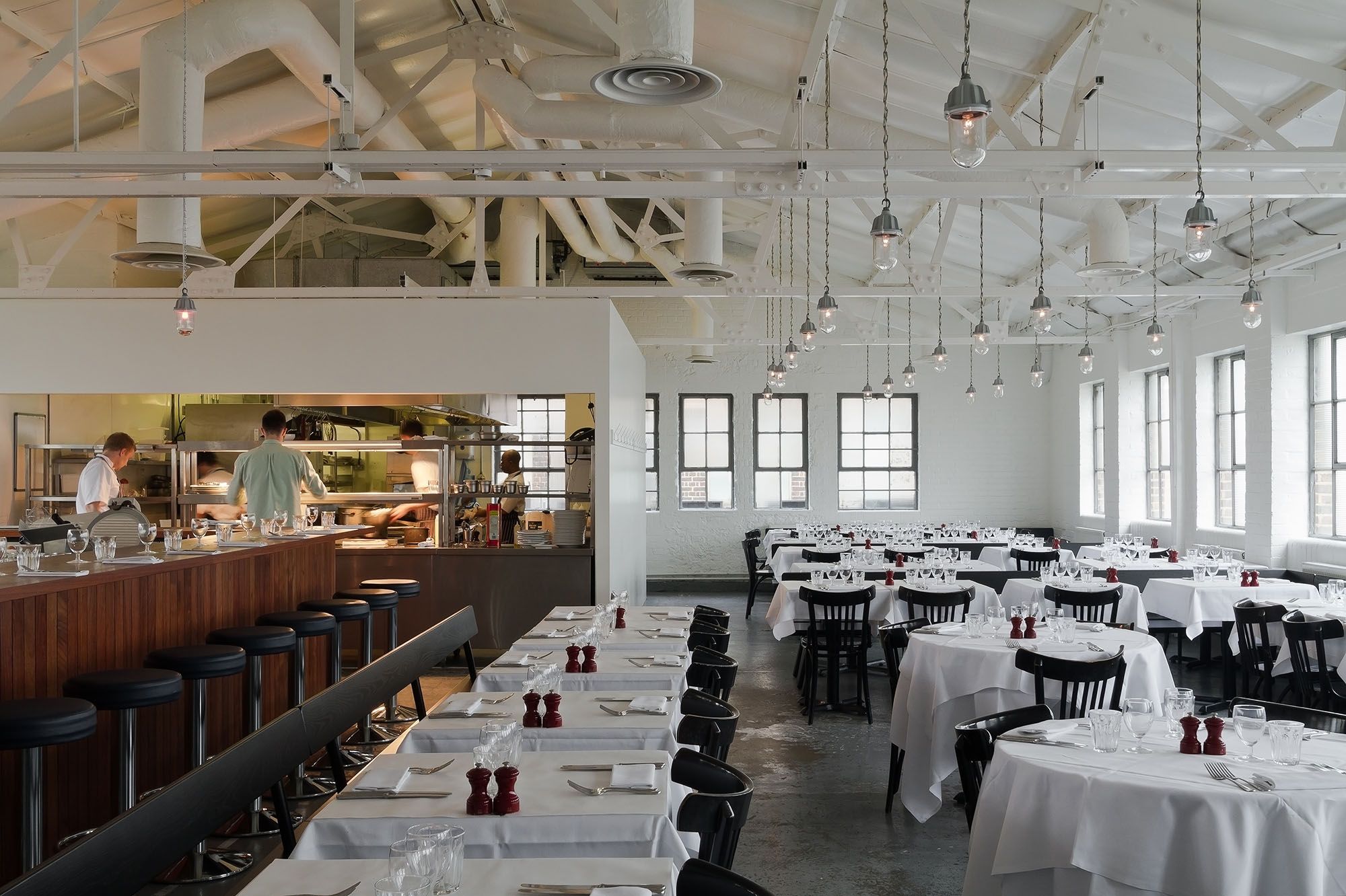 A bistro-style restaurant with white table cloths, dark wooden chairs, round tables, oendant lighting, and a wooden bar decked up with tall black leather stools, alongisde an open kitchen.