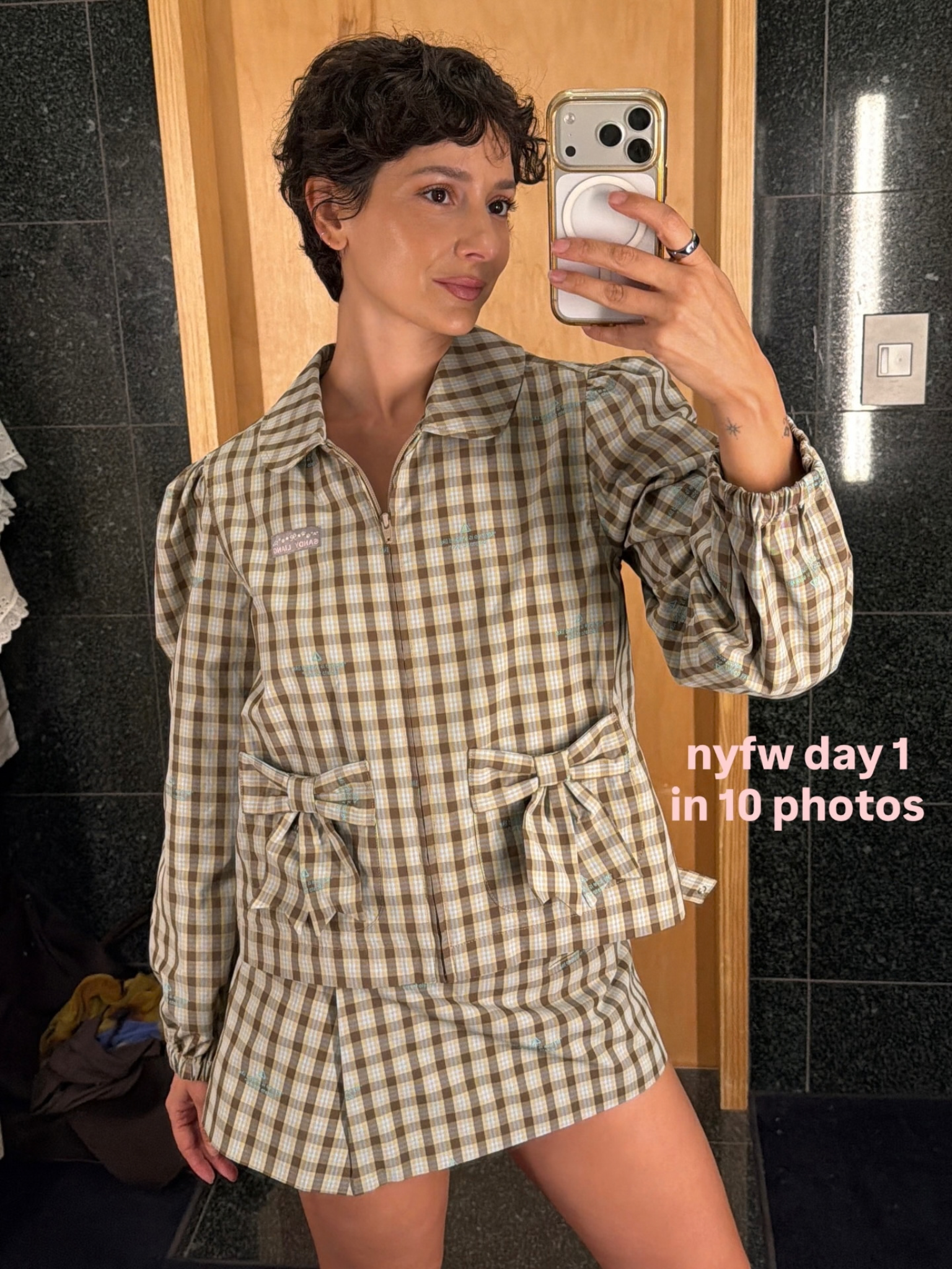 A woman in a checked shirt and skirt with a curly pixie haircut, taking a selfie in a mirror.