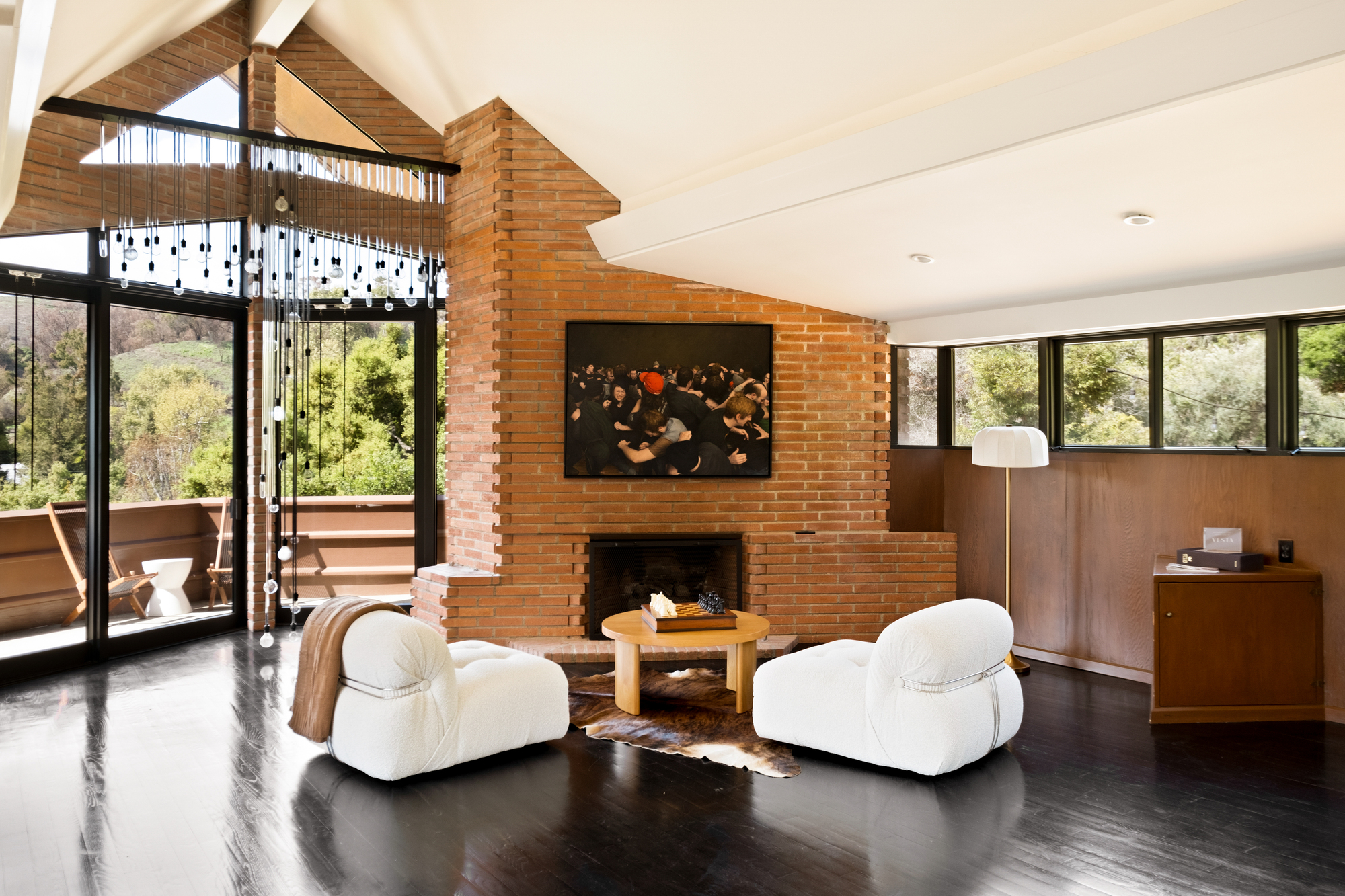 Image of an open concept living room with white ceilings, a large brick fireplace, and glossy black wooden floors. There are two white accent chairs, a cow hide rug, and a circular wooden coffee table positioned in front of the fireplace.
