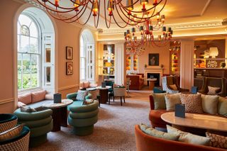 A vibrantly decorated hotel lobby features chubby, whimsical seating in green velvet and terracotta hues, orangey chandeliers, soft carpet, framed artworks, and lots of collectibles on its shelves.