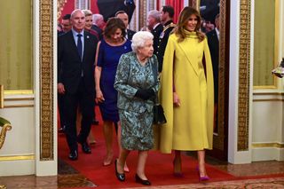 Queen Elizabeth walking next to Melania Trump wearing a yellow cape in Buckingham Palace