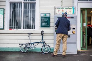 A man in beige trousers and a navy jacket stands in front of a railway ticket machine while his Brompton bicycle leans against the wall nearby