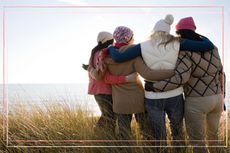 A group of four women looking out to sea and away from the camera with their arms around each other as they support their friend through early menopause