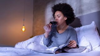 Woman drinking a glass of wine in a white bed wearing pajamas in a dim lit room