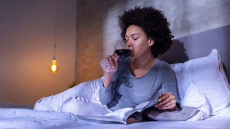 Woman drinking a glass of wine in a white bed wearing pajamas in a dim lit room