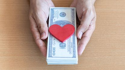 A pair of hands hold a stack of cash with a heart knickknack on top.