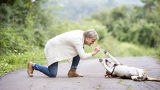 An older woman gives her dog a treat as it rolls over on a nature path.