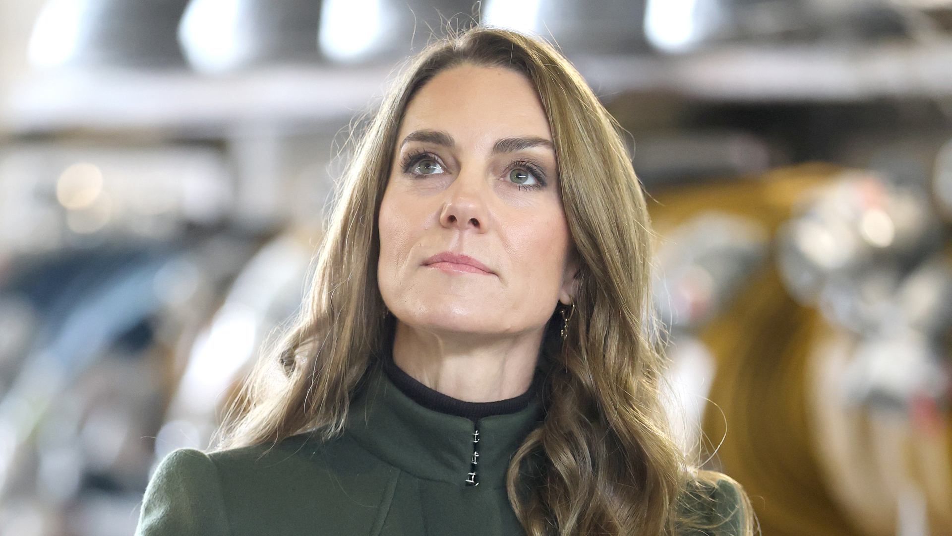 Princess Kate Middleton has long brown wavy hair with blonde highlights and looks to the sky while wearing a dark green coat