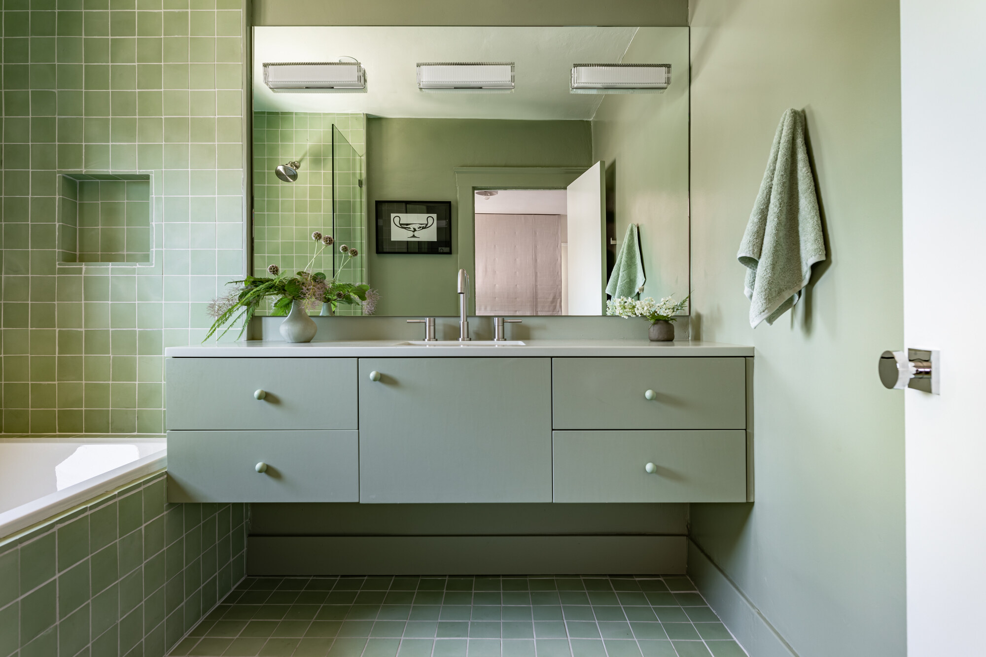 A sage green bathroom with sage green floating vanity, sage green zelliege tiles on the walls, floor, and built-in bath, sage green painted walls, sage green tiles, and a vase of flowers styled on the counter
