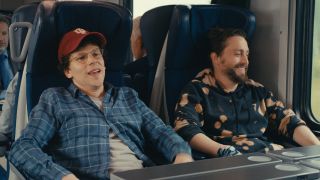 From left to right: Jesse Eisenberg and Kieran Culkin smiling on a train.