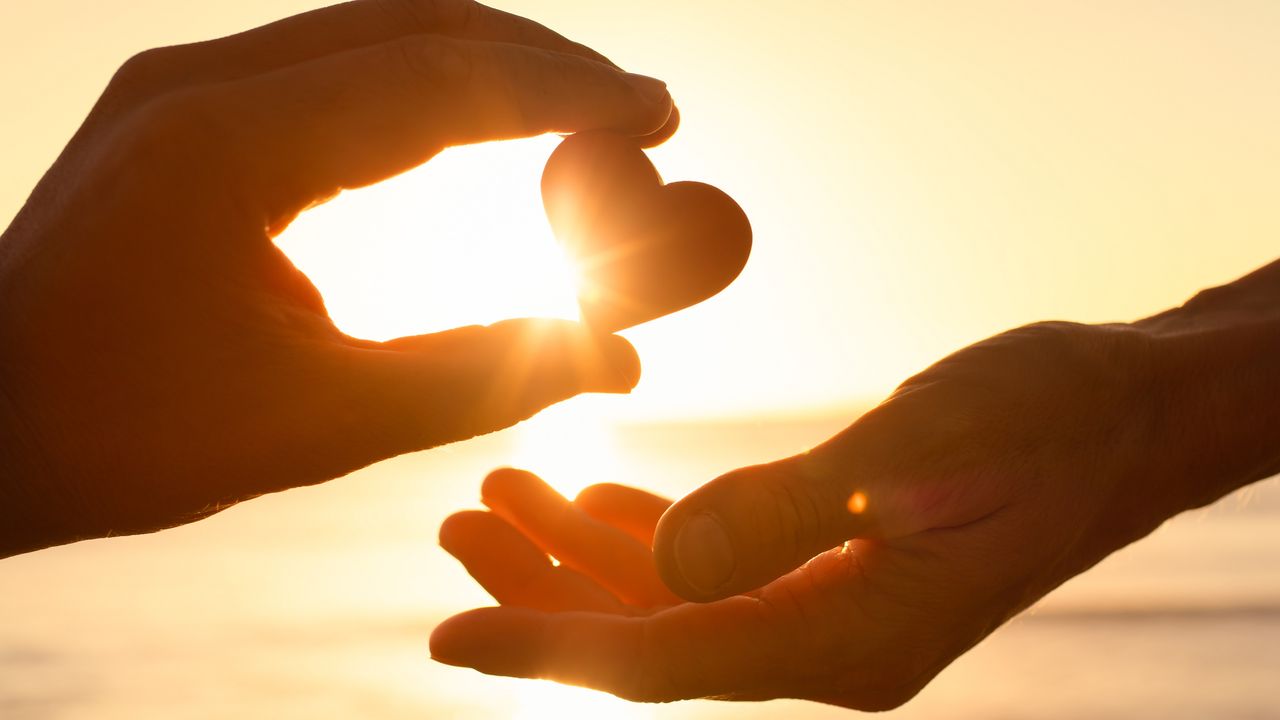 The sun shines behind a paper heart as one person gives the heart to another, only their hands showing.