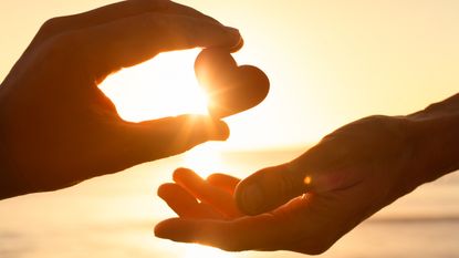 The sun shines behind a paper heart as one person gives the heart to another, only their hands showing.