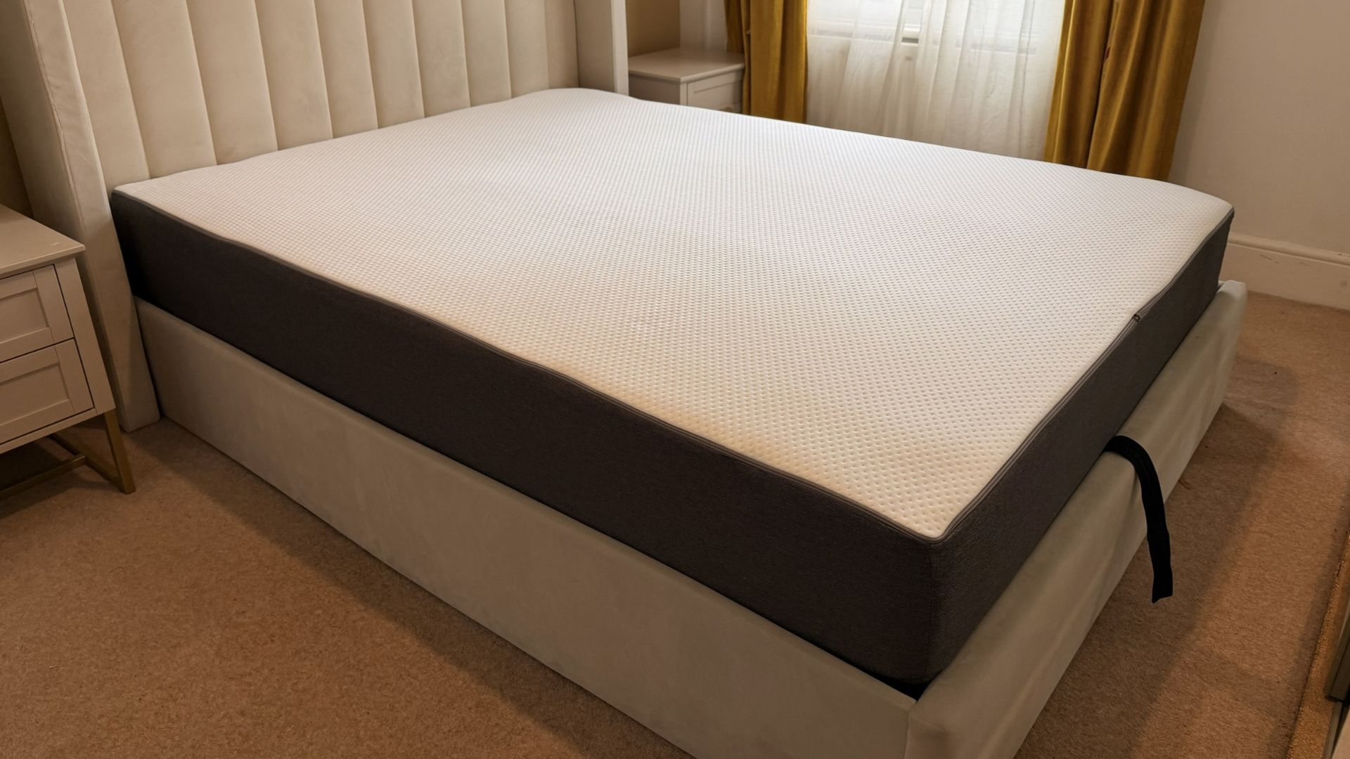 The image shows a grey and white mattress on a grey fabric bed frame, set within a neutrally decorated bedroom