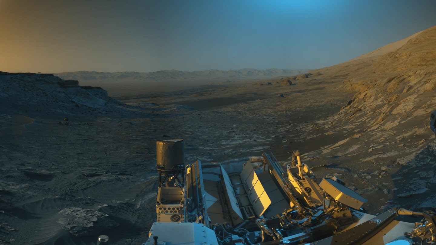Martian panorama showing the view back down Mount Sharp at the center of the image and rounded hills in the distance.