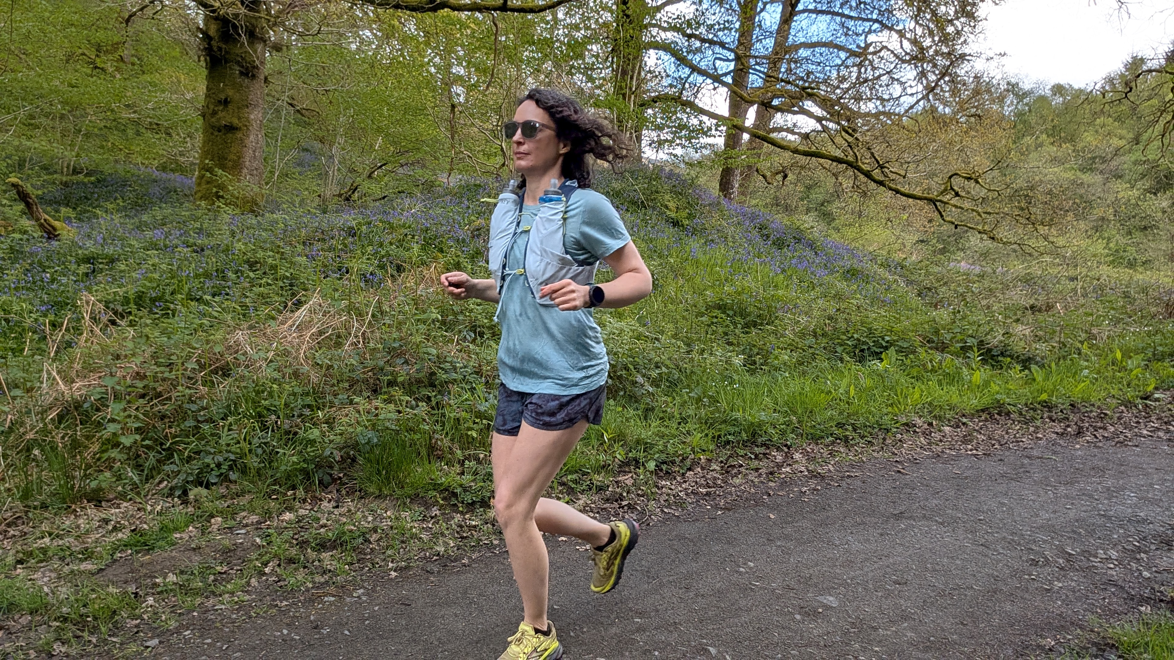 A woman running on a forest trail wearing a light blue T-shirt and Osprey Dyna running vest