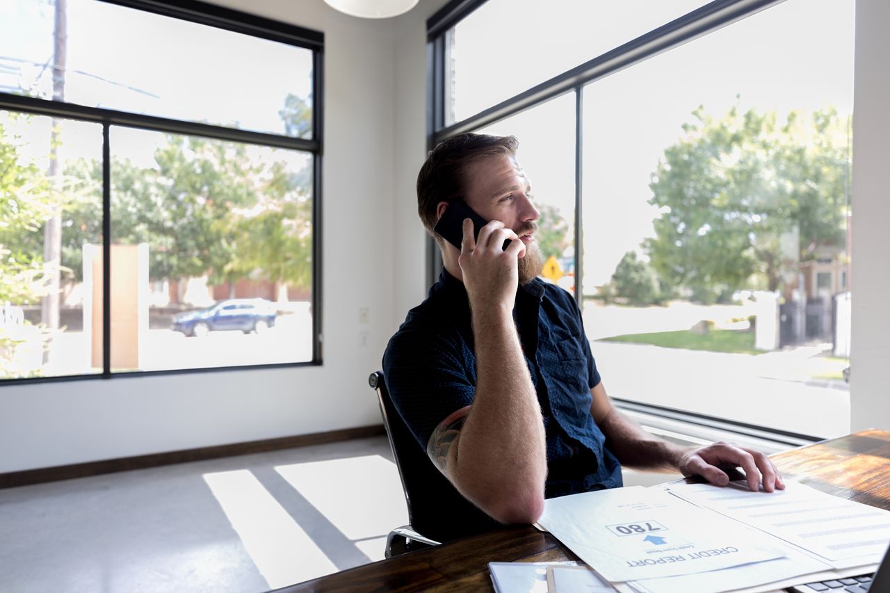 Businessman gazes through a window while he takes a phone call. A credit report is on his desk.