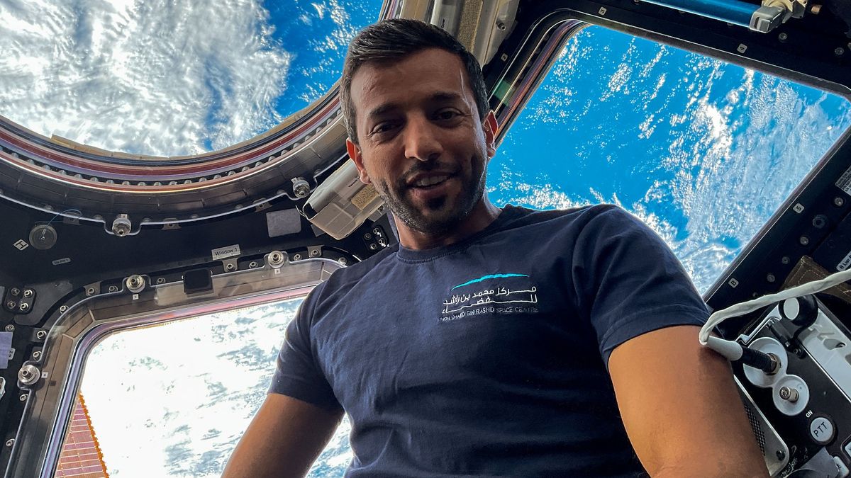 sultan al-neyadi in front of three large windows in a space station. behind is the earth with clouds on the surface