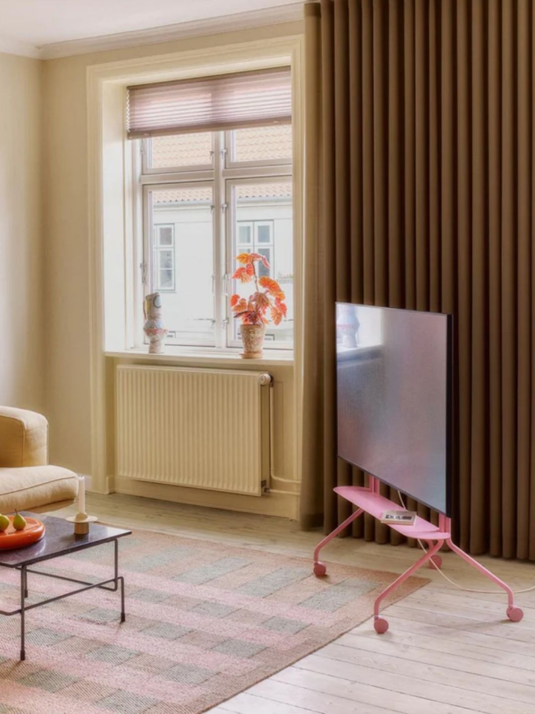 A beige/light yellow room with brown curtains and a pink plaid flatweave rug with a TV that's on a light pink rolling TV stand.