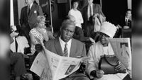 Black and white photograph of Albert Luthuli sat with a newspaper in his hands, as he looks directly into the camera. His wife is sat beside him.