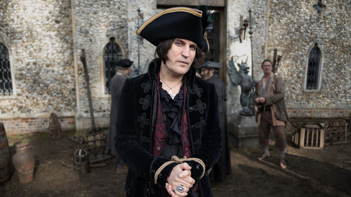 Noel Fielding in a black hat and jacket with his hands tied together in The Completely Made-Up Adventures of Dick Turpin