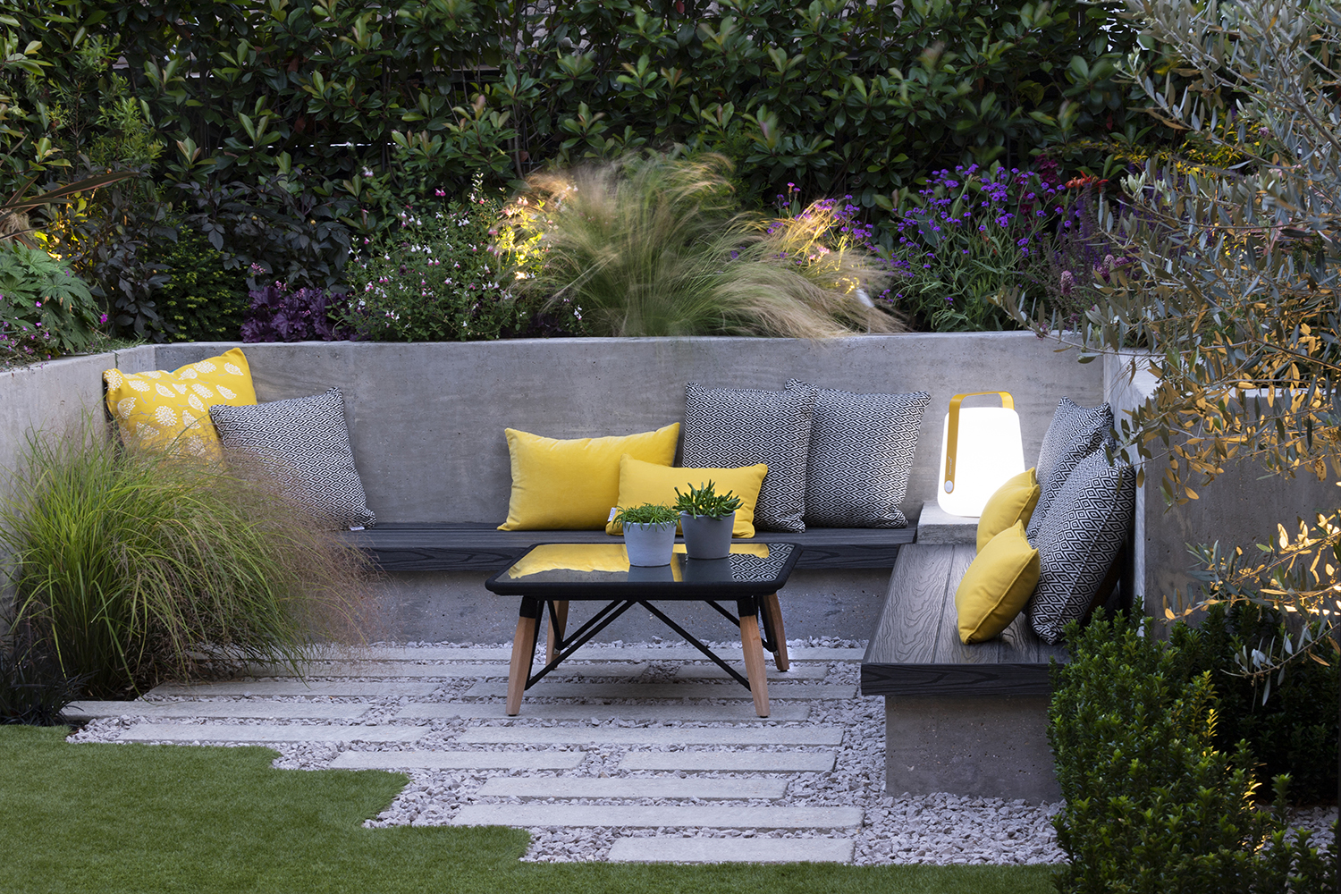 a sunken seating area in a backyard, with gravel and paver floors, a lawn, a built-in bench seat with yellow and gray cushions, and a square coffee table