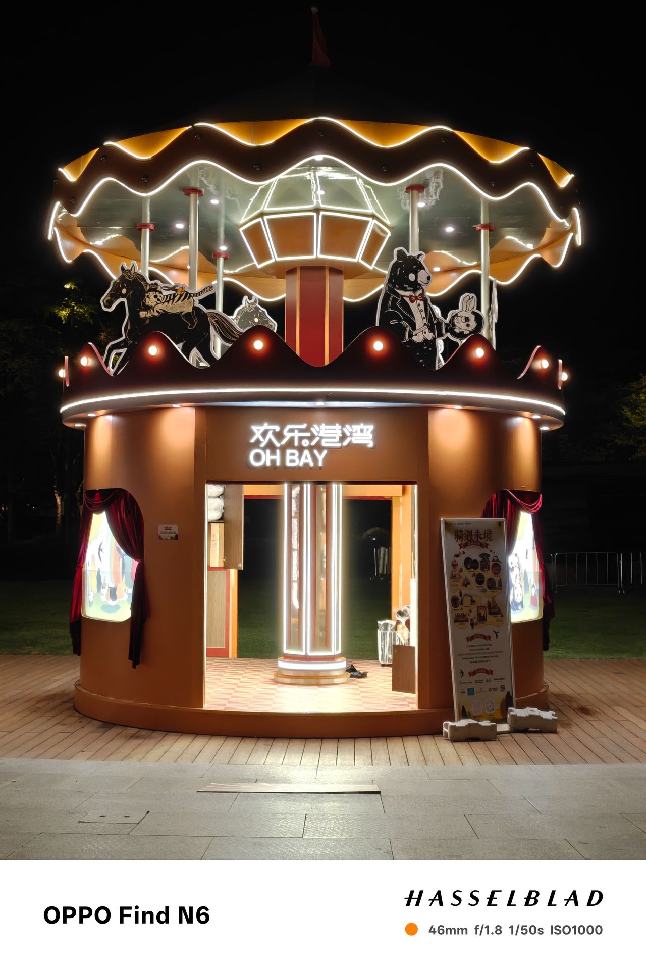 A medium shot of a whimsical, carousel-themed ticket booth or kiosk at night. The structure is glowing with warm yellow lights and features decorative cutouts of a horse and a bear. The words "OH BAY" are illuminated in white neon above the entrance.