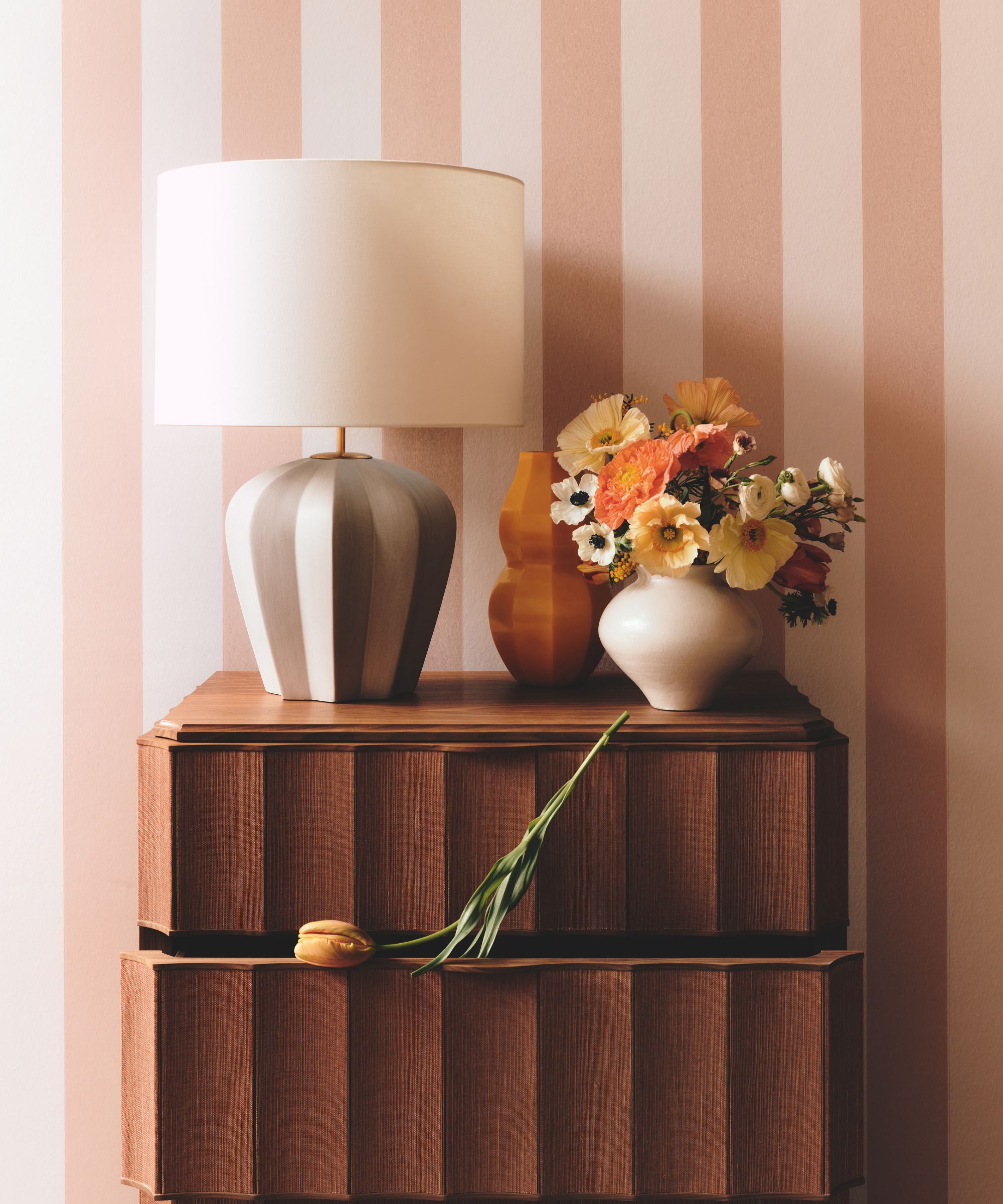 A mid-century modern wooden dresser against pink-and-white striped wallpaper. A grey fluted lamp and a white vase of orange and yellow poppies sit on top, with a single tulip draped over an open drawer.