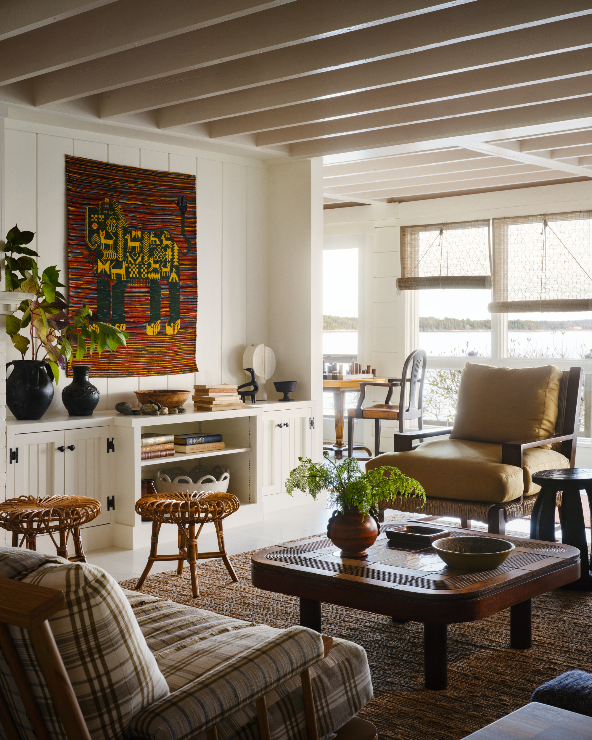 Image of a white, wood-panelled living room with a red tapestry on the wall, a jute rug, a yellow accent chair, a plaid sofa, and a wooden coffee table.