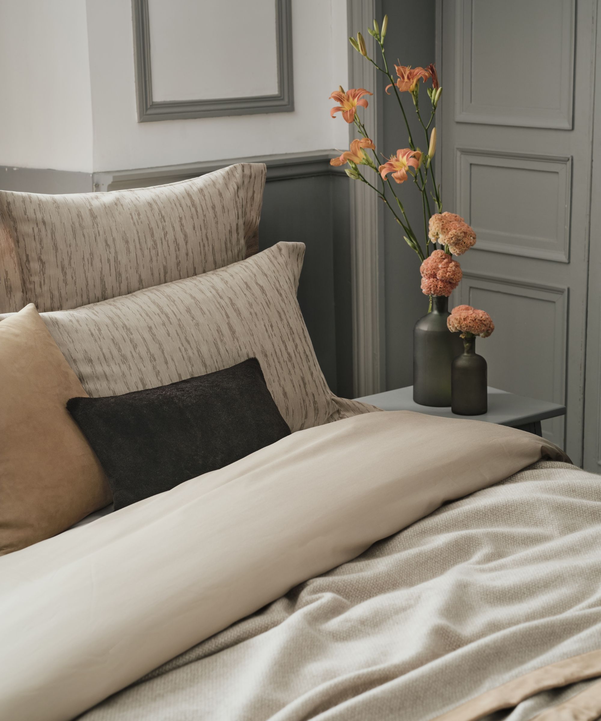 A close-up of a corner of a bed with beige bedding, cushions and pillows in a neutral grey and white panelled bedroom with pink flowers on a grey bedside table beside it