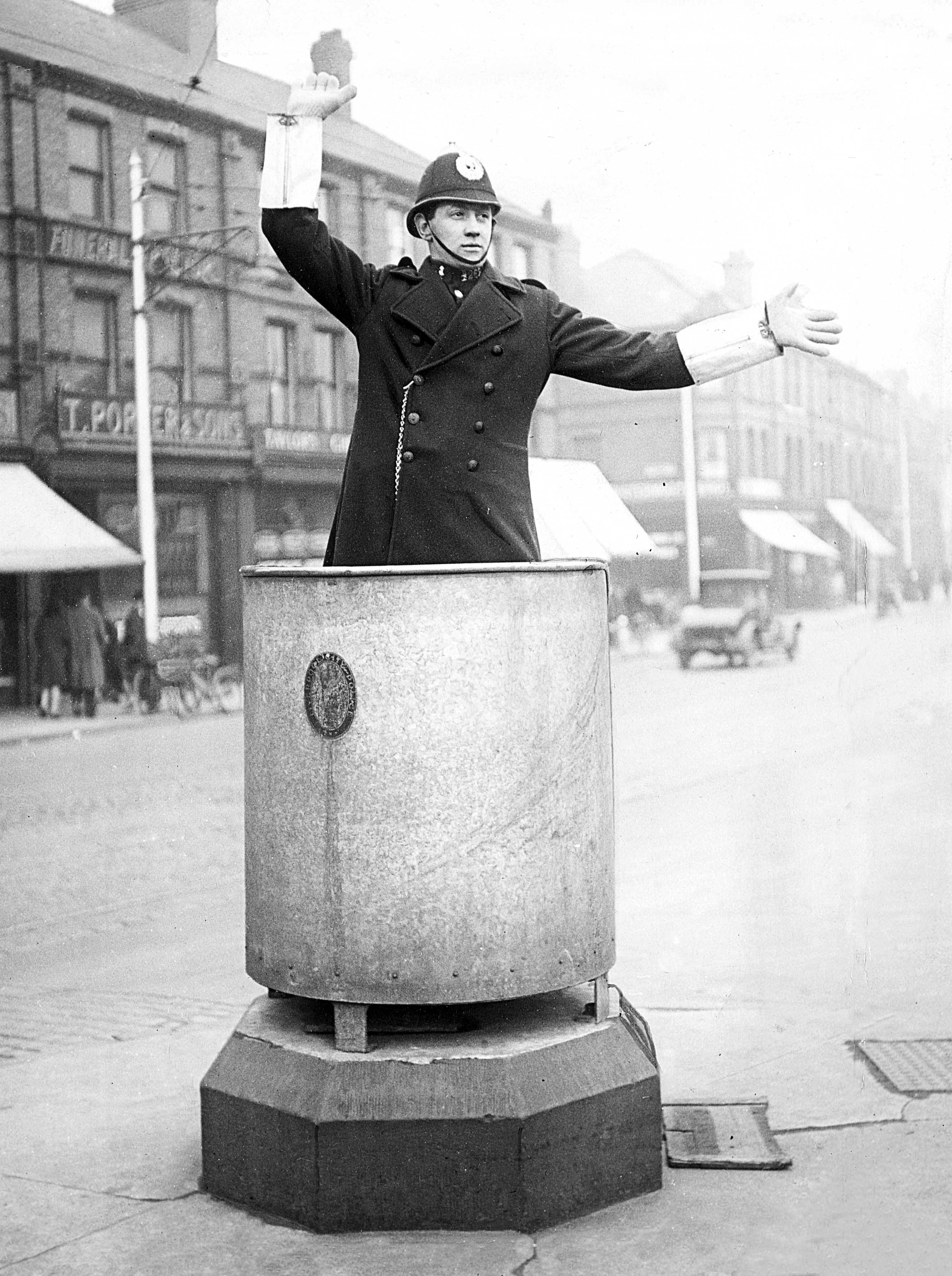 A police officer in a helmet and greatcoat stands inside a large metal drum on a plinth in the middle of a Liverpool street, arms outstretched to direct traffic, with shops and a car behind him.