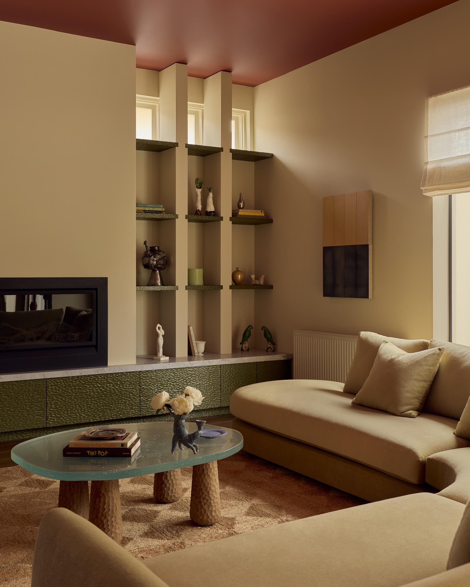 modern living room with beige corner sofa, jute rug with check detail, glass coffee table with chiselled timber legs, built-in shelving next to a fireplace, green drawers and red ceiling