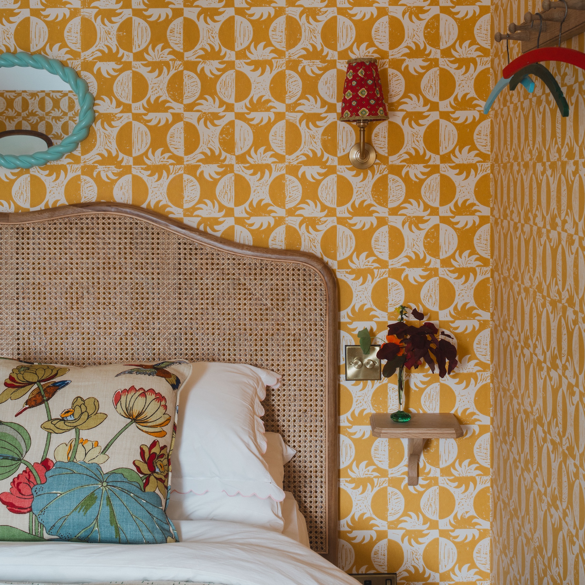 bedroom with patterned yellow wallpaper, double bed with wicker headboard and colourful cushions, blue frame mirror on wall and wall lights with pleated lampshades