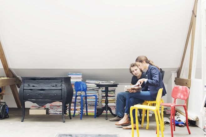 A man and woman sitting in a work room with items stackd up against the wall. 