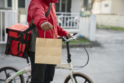 A delivery person on a bike carries a food order in a brown bag.