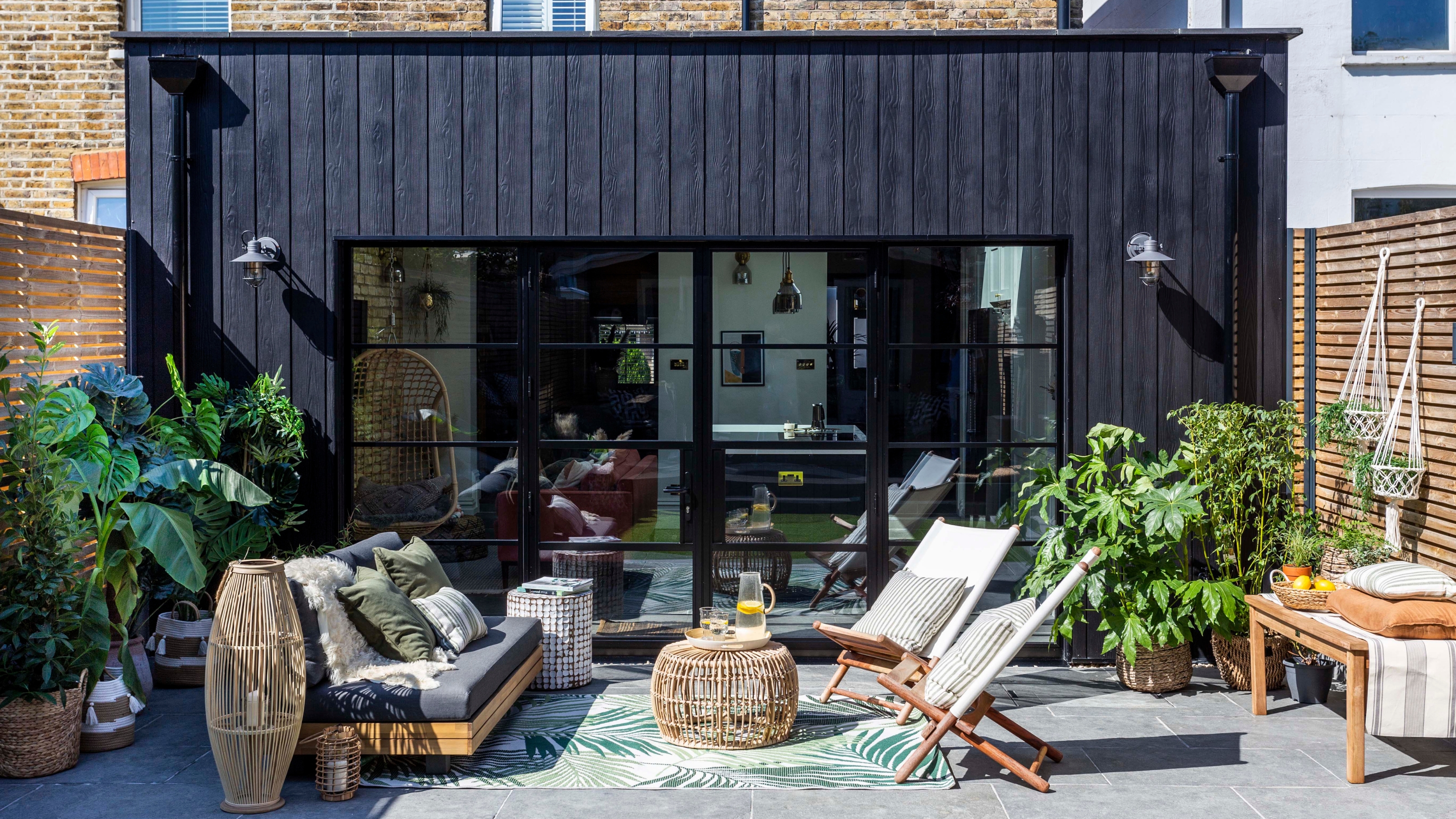 Image of the exterior of a home and patio. The house has black fibre cement cladding and wooden fencing on either side of the patio. There is an outdoor seating setup with a loveseat and two sling chairs. 