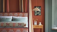 Wall decor for bedroom, featuring terracotta colored walls with gray-blue window frames and shutters and geometric print wall art, headboard and soft furnishings.