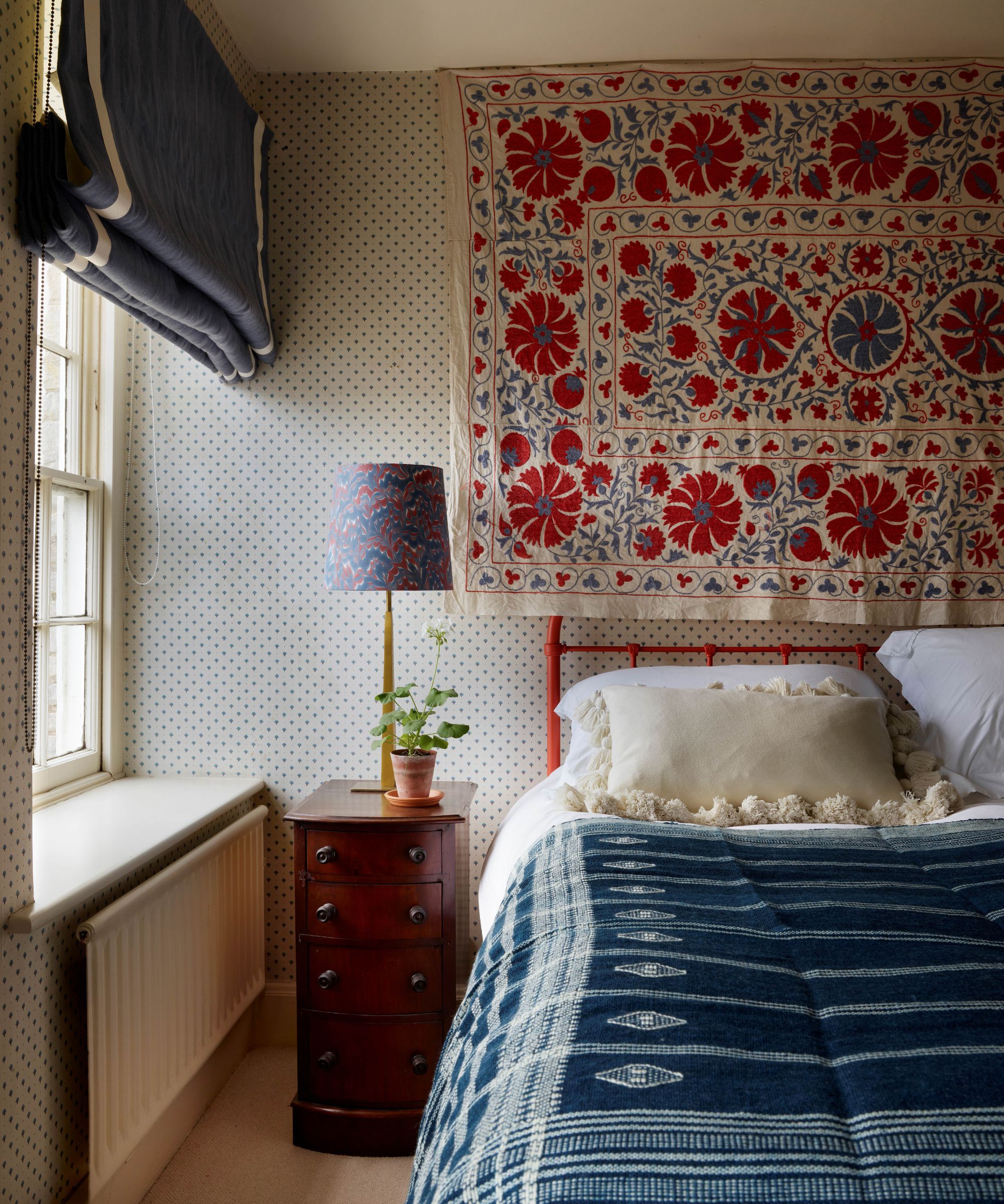 An eclectic bedroom featuring a red bed frame, a blue patterned bedspread, and a large red and white floral textile hanging on the wall above the headboard