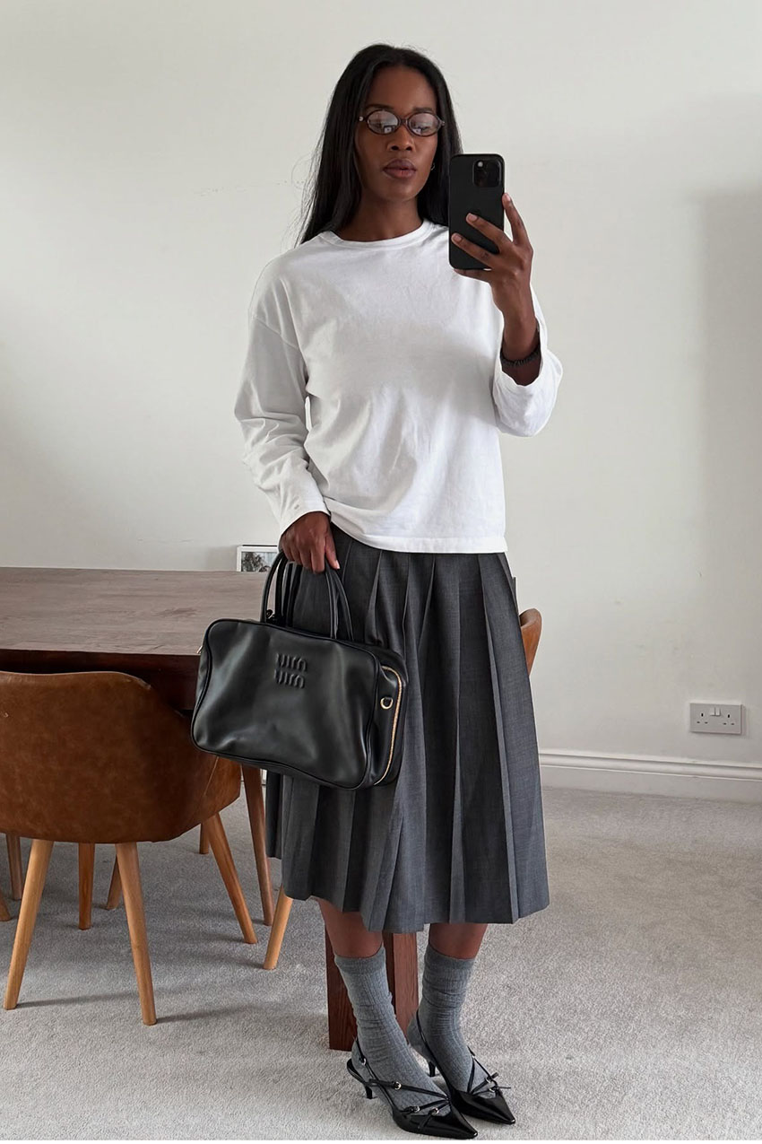 A woman wearing a white long-sleeve shirt, gray skirt, gray socks, black heels, and a black bag.